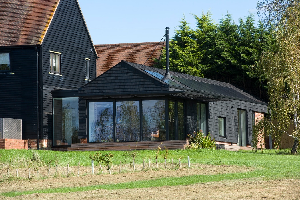 Modern black-clad house extension with large glass windows, connected to an older brick and timber house, surrounded by grass and trees.