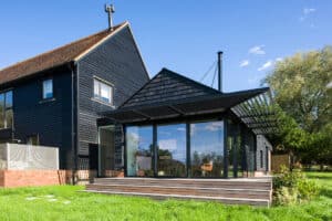 Modern black timber house with large glass doors, a wooden deck, and a sloped roof, situated on a grassy lawn under a clear blue sky.