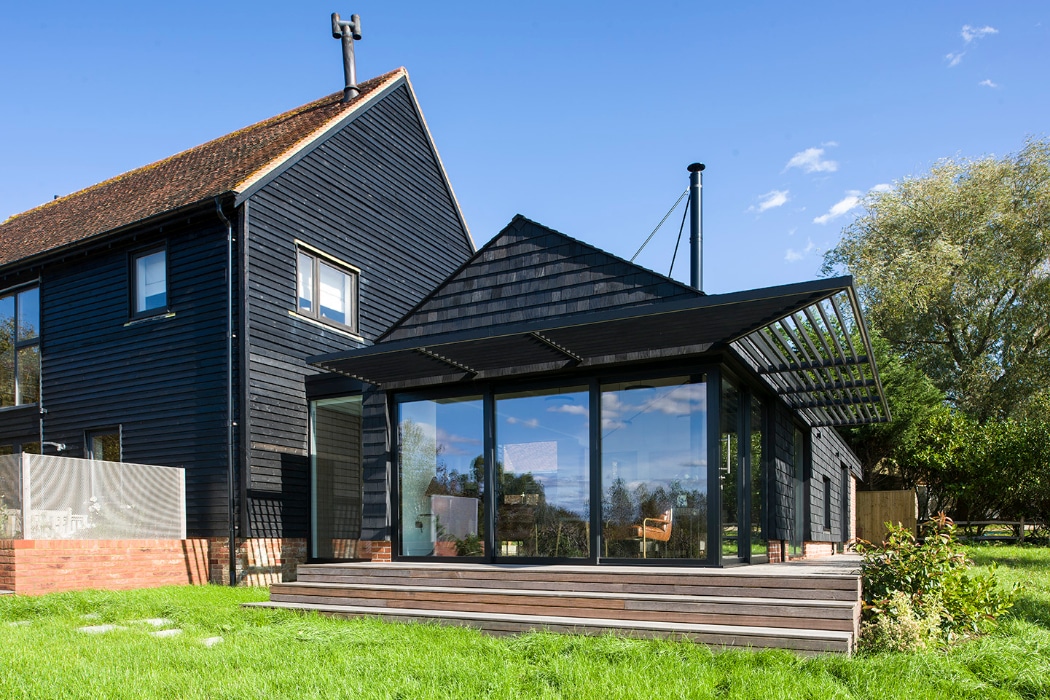 Modern black timber house with large glass doors, a wooden deck, and a sloped roof, situated on a grassy lawn under a clear blue sky.