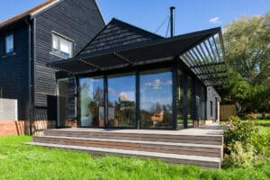 Modern glass-walled home extension with a black frame and louvered roof, attached to a dark-paneled house, surrounded by green grass and trees.