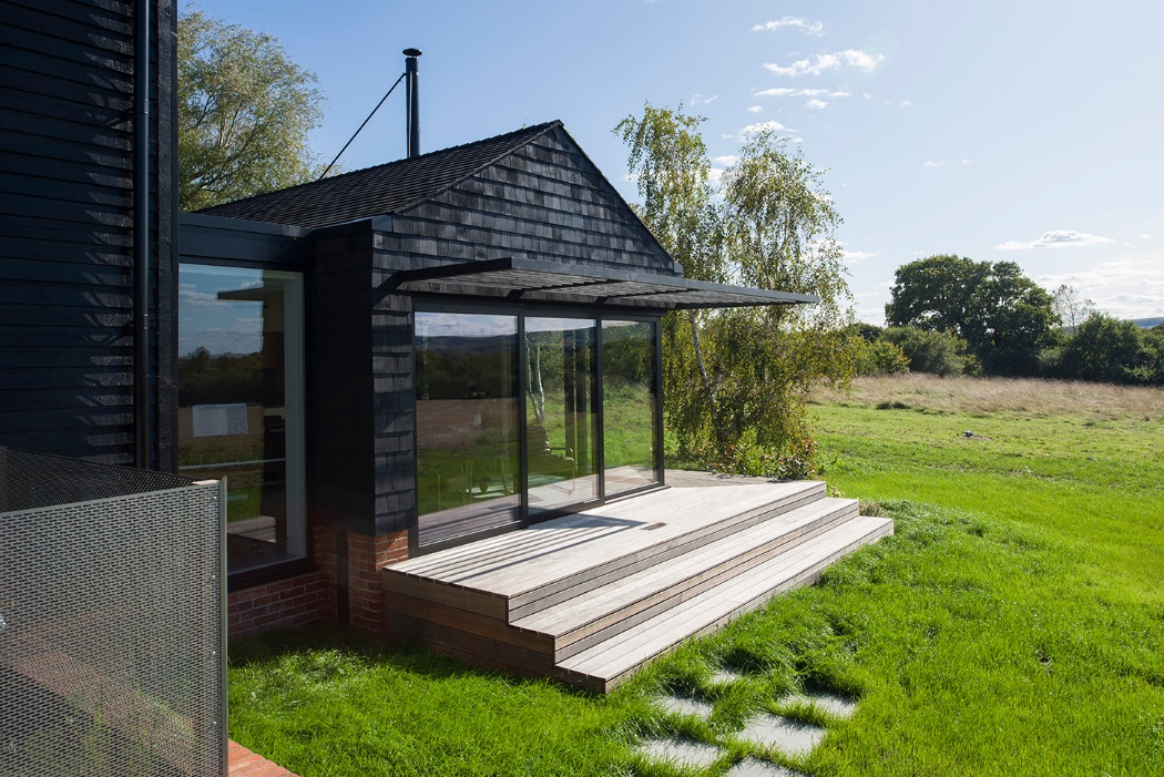 A modern house with dark wood siding features a glass-walled extension, a wooden deck with steps, and overlooks a grassy yard under a clear sky.
