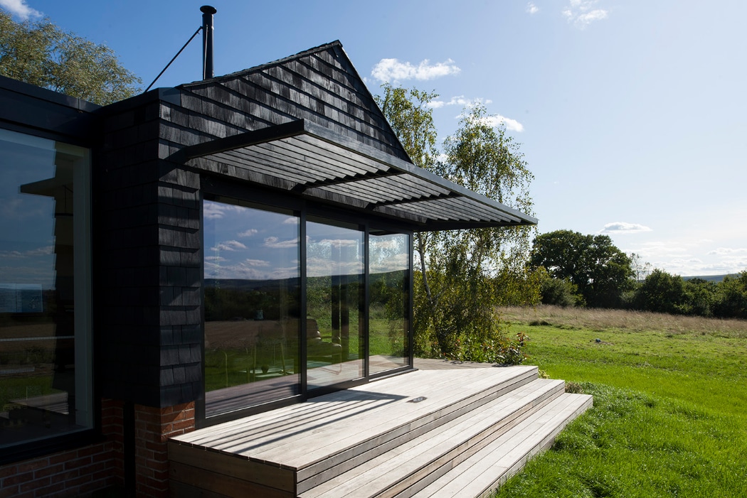 A modern house with large glass sliding doors, a wooden deck with steps, and a black slatted awning, overlooking a grassy field and trees under a blue sky.