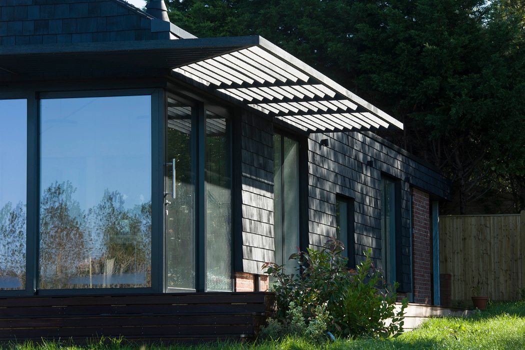 Modern house with large glass windows, dark shingle siding, and a slatted overhang, set against greenery and a wooden fence in the background.