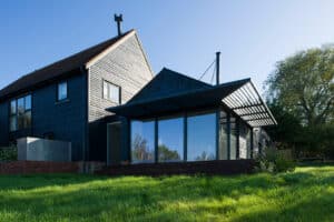 Modern black house with large glass windows and wooden exterior, situated on a grassy lawn with clear blue sky in the background.