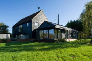 A modern black house with a pitched roof and a large glass-walled extension, situated on a grassy lawn under a clear blue sky.