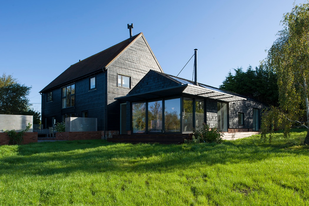 A modern black house with a pitched roof and a large glass-walled extension, situated on a grassy lawn under a clear blue sky.