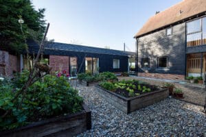 Modern courtyard garden with raised wooden planters filled with plants and gravel ground cover, surrounded by black timber and brick buildings with large windows.