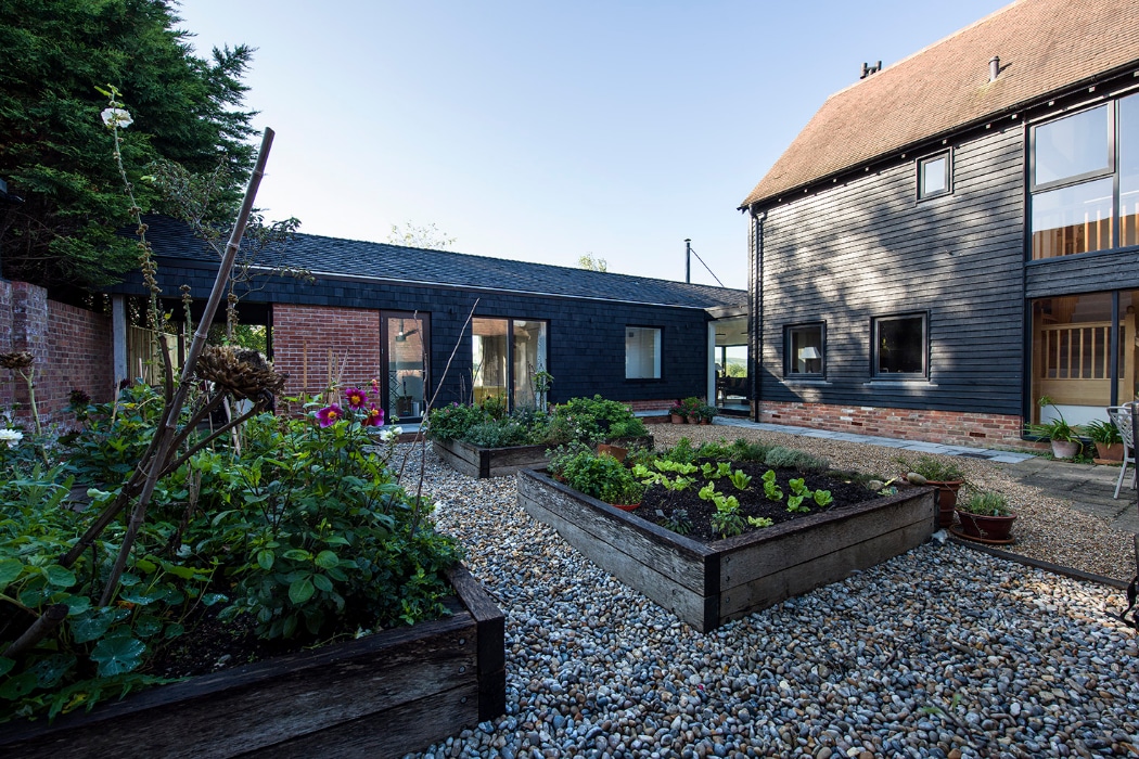 Modern courtyard garden with raised wooden planters filled with plants and gravel ground cover, surrounded by black timber and brick buildings with large windows.