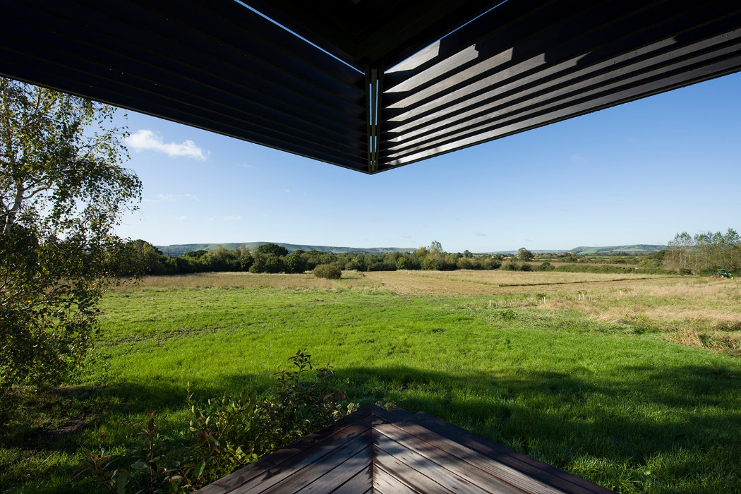 View of a grassy field and distant hills seen from beneath the shaded corner of a modern building with wooden decking in the foreground.