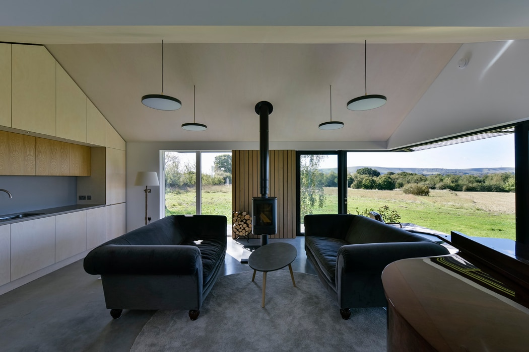 Modern living room with two dark sofas facing a wood stove, large windows with views of fields, minimalist kitchen on the left, and suspended circular lights.