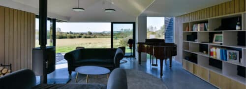 Modern living room with large windows, black sofas, a round coffee table, a grand piano, bookshelf, and wood-burning stove, overlooking a grassy landscape and distant hills.
