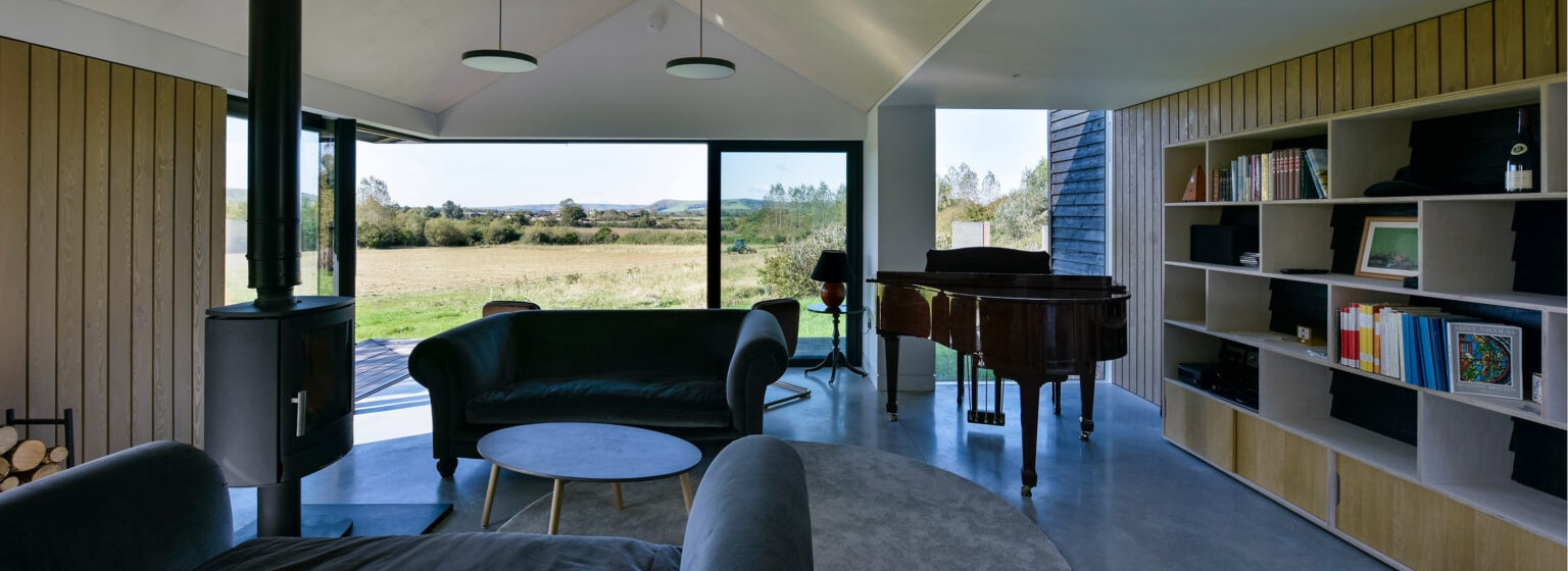 Modern living room with large windows, black sofas, a round coffee table, a grand piano, bookshelf, and wood-burning stove, overlooking a grassy landscape and distant hills.
