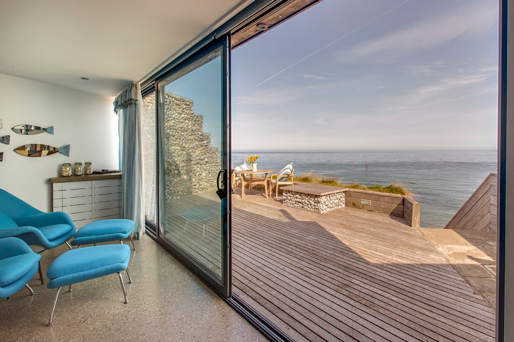 Modern living room with blue chairs and large glass doors opening to a wooden deck with outdoor seating and a view of the ocean.