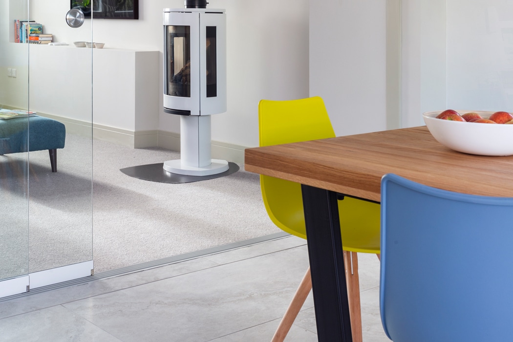 A modern dining area with a wooden table, yellow and blue chairs, and a bowl of apples; a single sliding door opens to reveal a white stove and living space in the background.
