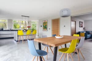 Modern open-plan kitchen and dining area with colorful chairs, wooden table, and large windows; a single sliding door connects to the living room visible in the background.