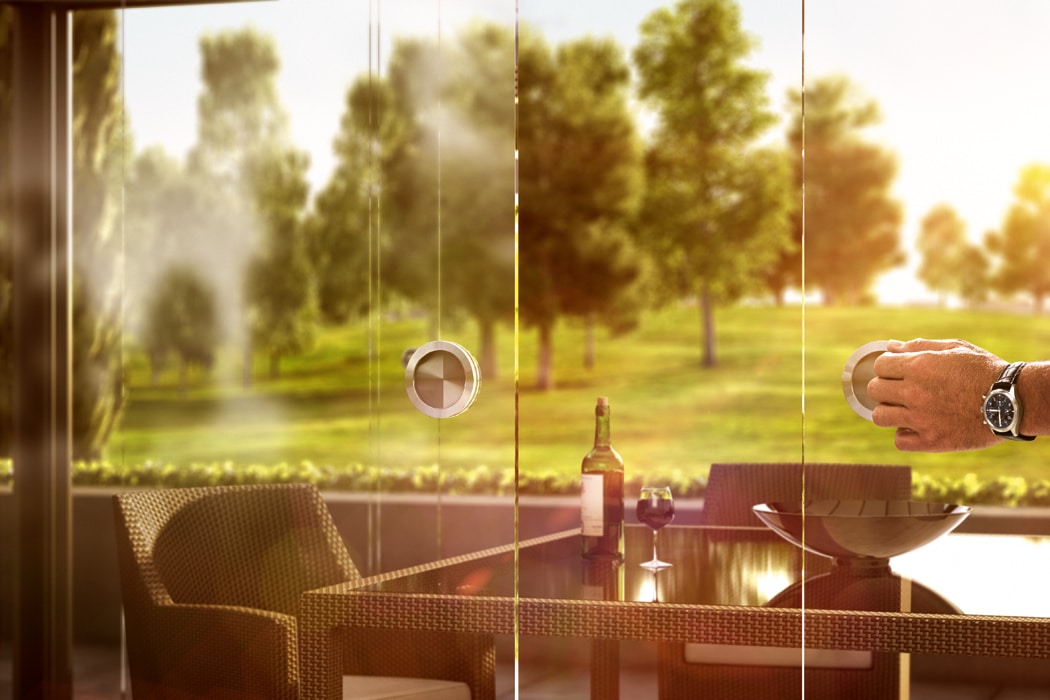 A person opens a single sliding door to an outdoor patio with wicker chairs, a table, a wine bottle, and a glass. Trees and grass are visible in the background.