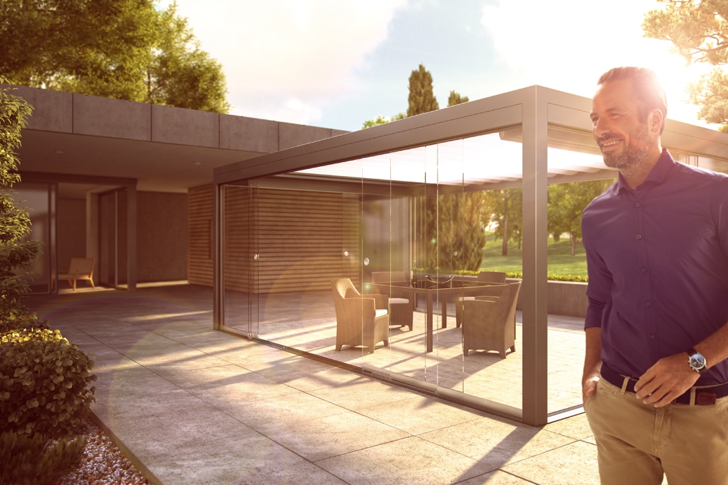 A man stands outside a modern house with a glass-enclosed patio, sunlight streaming through the single sliding door and highlighting the patio furniture inside.
