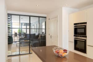 Modern kitchen with built-in ovens, white cabinets, and a fruit bowl on the island; heritage doors with glass panels reveal a living area and patio beyond.