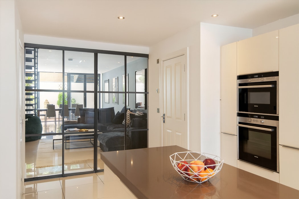 Modern kitchen with built-in ovens, white cabinets, and a fruit bowl on the island; heritage doors with glass panels reveal a living area and patio beyond.