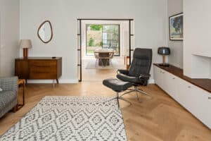 Modern living room with herringbone wood floors, patterned rug, black lounge chair, heritage doors, mid-century furniture, and a view into a dining area with large windows to a garden.