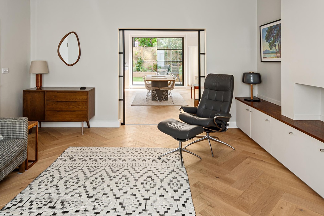 Modern living room with herringbone wood floors, patterned rug, black lounge chair, heritage doors, mid-century furniture, and a view into a dining area with large windows to a garden.