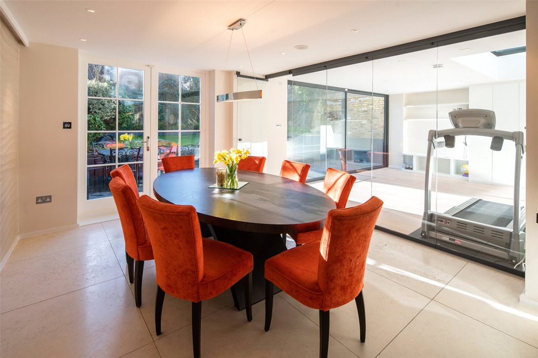 A dining room with eight orange chairs around a dark oval table, a vase of yellow flowers, and a treadmill visible in an adjacent glass-walled room with slide and turn doors.