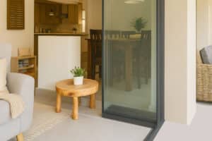 A modern living space with a wooden table and potted plant near elegant slide and turn doors, with a dining area and kitchen visible in the background.