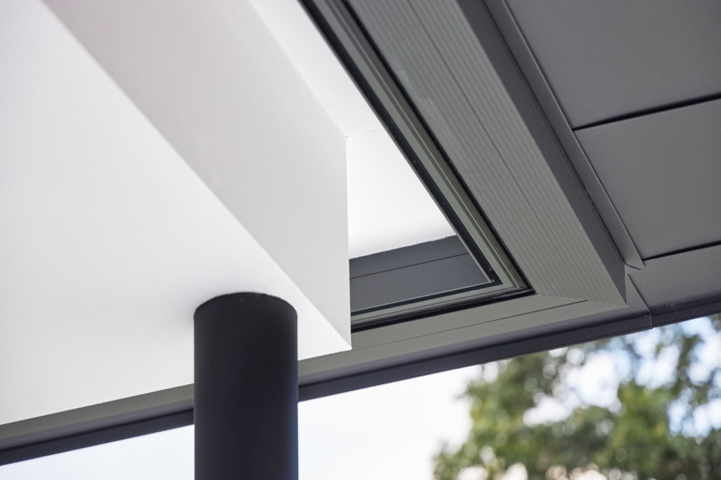 A close-up view of a modern building ceiling corner with a black cylindrical column and an opening, showing part of the sky and tree in the background.