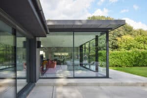Modern house with large floor-to-ceiling glass windows and patio, showing a minimalist interior and surrounding green lawn and trees.