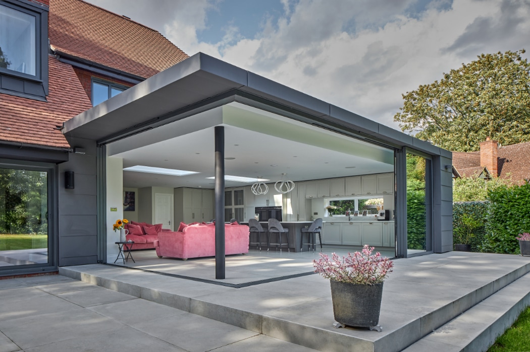 Modern house with large glass sliding doors open to a patio, showing a spacious kitchen and living area with pink sofas and contemporary lighting inside.