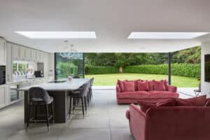 Modern open-plan kitchen and living area with large island, red sofas, skylights, and floor-to-ceiling glass doors overlooking a green garden.
