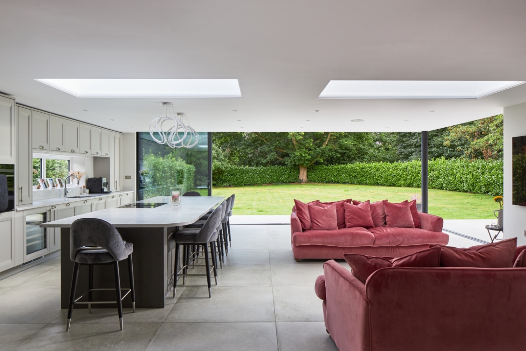 Modern open-plan kitchen and living area with large island, red sofas, skylights, and floor-to-ceiling glass doors overlooking a green garden.