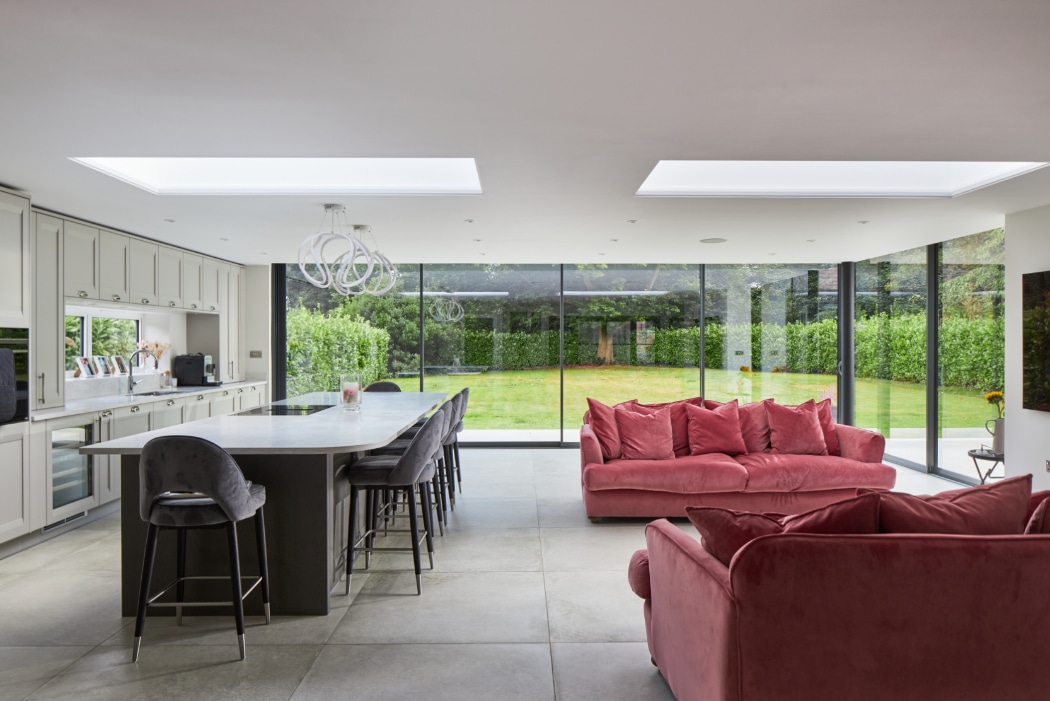 Modern open-plan kitchen and living area with large windows, white cabinetry, an island with barstools, and red velvet sofas, overlooking a green garden.