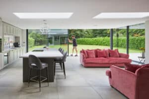 Modern open-plan kitchen and living area with large windows, pink sofas, and a person sliding open a glass door to a green garden outside.