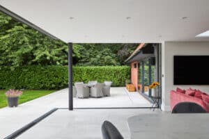 Modern open-plan living space with sliding glass doors leading to a patio featuring an outdoor dining set, potted plant, and garden hedge.
