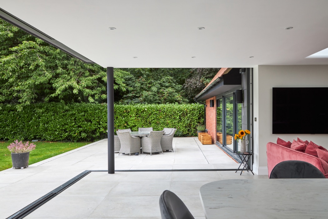 Modern open-plan living space with sliding glass doors leading to a patio featuring an outdoor dining set, potted plant, and garden hedge.