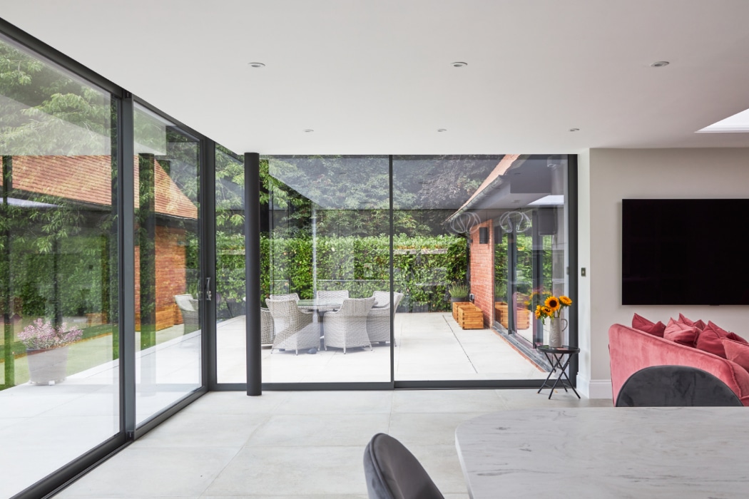 Modern open-plan living area with large glass walls looking out onto a patio with outdoor dining furniture and green hedges.