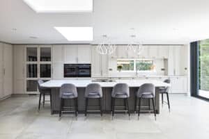 Modern kitchen with a large island featuring five gray barstools, pendant lights overhead, white cabinets, and a view of greenery through large windows.