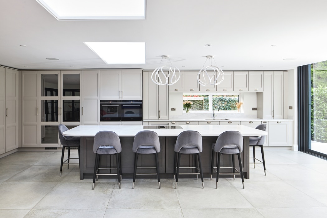 Modern kitchen with a large island featuring five gray barstools, pendant lights overhead, white cabinets, and a view of greenery through large windows.