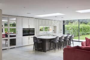 Modern kitchen with white cabinets, a large island with gray barstools, pendant lights, and floor-to-ceiling windows overlooking a green garden. Red seating is visible in the foreground.