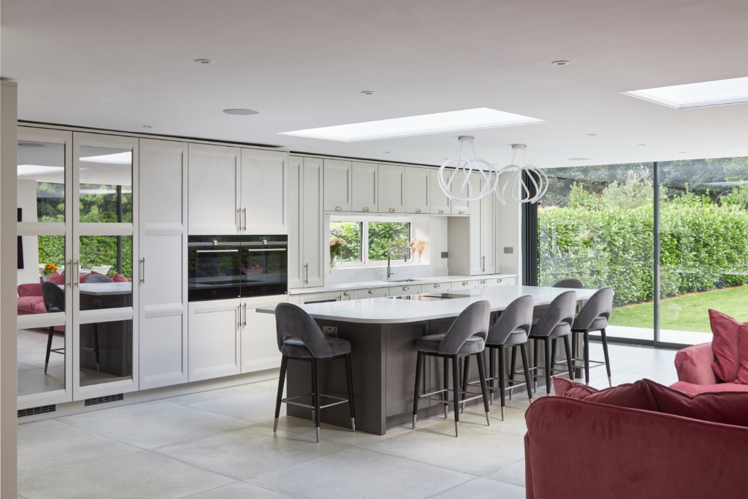 Modern kitchen with white cabinets, a large island with gray barstools, pendant lights, and floor-to-ceiling windows overlooking a green garden. Red seating is visible in the foreground.
