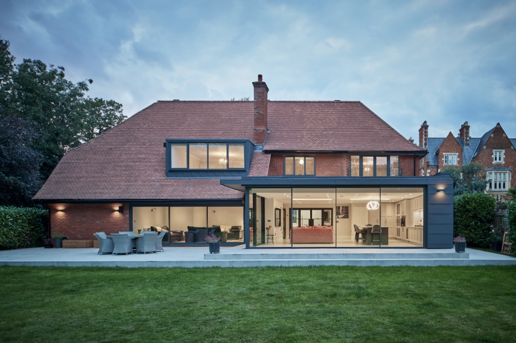 Modern two-story house with large glass windows, red-tiled roof, and spacious patio area opening onto a green lawn; neighboring houses in the background.
