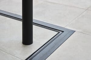 A black vertical pole intersects beige floor tiles with a black metal groove forming a right angle around the base.