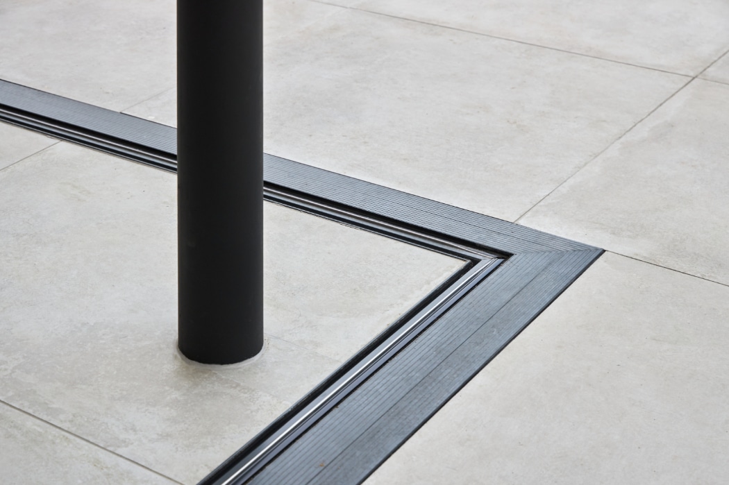 A black vertical pole intersects beige floor tiles with a black metal groove forming a right angle around the base.