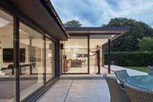 Modern house with large glass windows and sliding doors, showing an illuminated interior and a patio with outdoor furniture; a person is visible inside, appearing slightly blurred in motion.