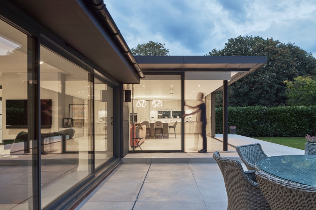 Modern house with large glass windows and sliding doors, showing an illuminated interior and a patio with outdoor furniture; a person is visible inside, appearing slightly blurred in motion.