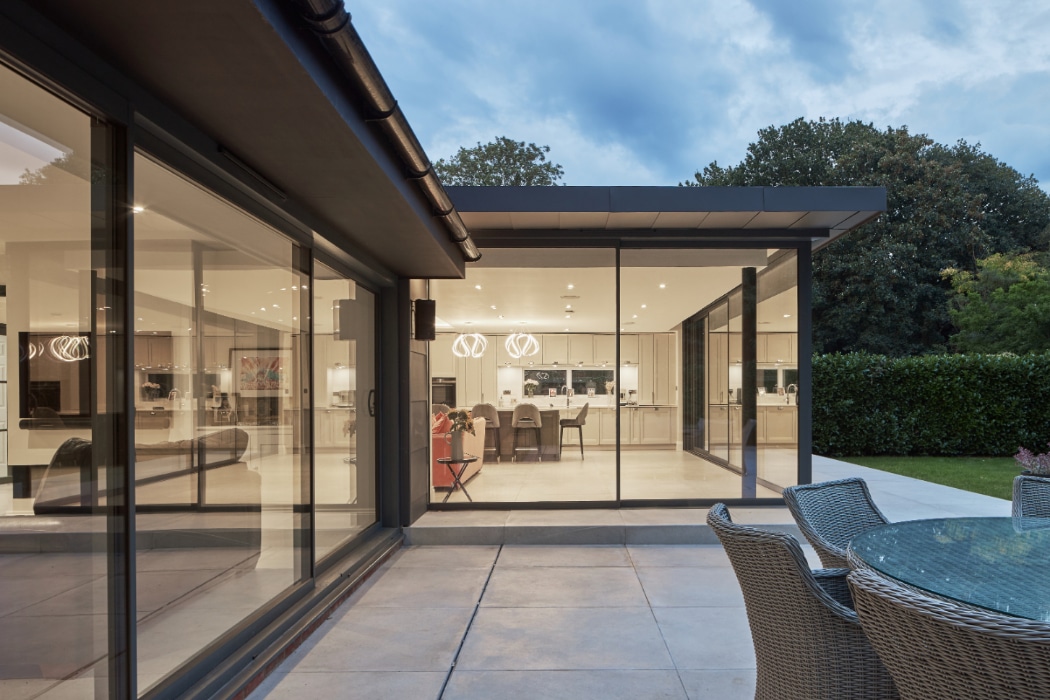 Modern house with large glass sliding doors and windows, outdoor patio with a round glass table and wicker chairs, interior visible with contemporary lighting.