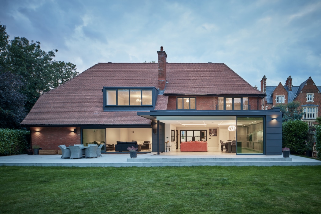 A large modern house with brick walls, large windows, a glass extension, and an outdoor patio set, viewed from the back garden at dusk.