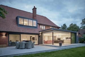 Modern house with large glass doors opening to a patio with outdoor dining furniture, showcasing an illuminated open-plan kitchen and living area inside.