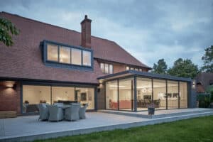 Modern house with large glass extension, patio dining set, and spacious backyard lawn under a cloudy sky.